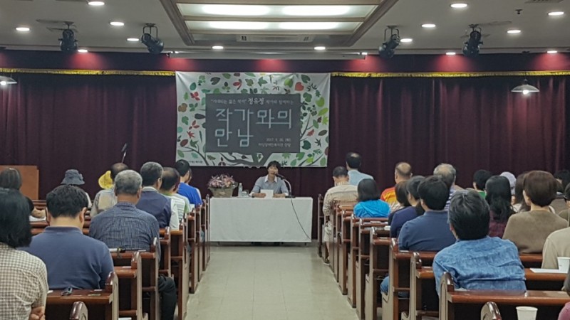 복지관 강당 앞에서 정유정 작가가 테이블 뒤에 앉아서 청중들에게 이야기를 하고 있는 모습과 정유정 작가의 말을 경청하고 있는 관중들의 모습