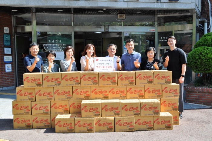 복지관 현관에서 후원품 라면 45박스와 서울강남우체국 임직원 6명과 복지관 국장, 전략기획팀장이 함께 찍은 단체사진